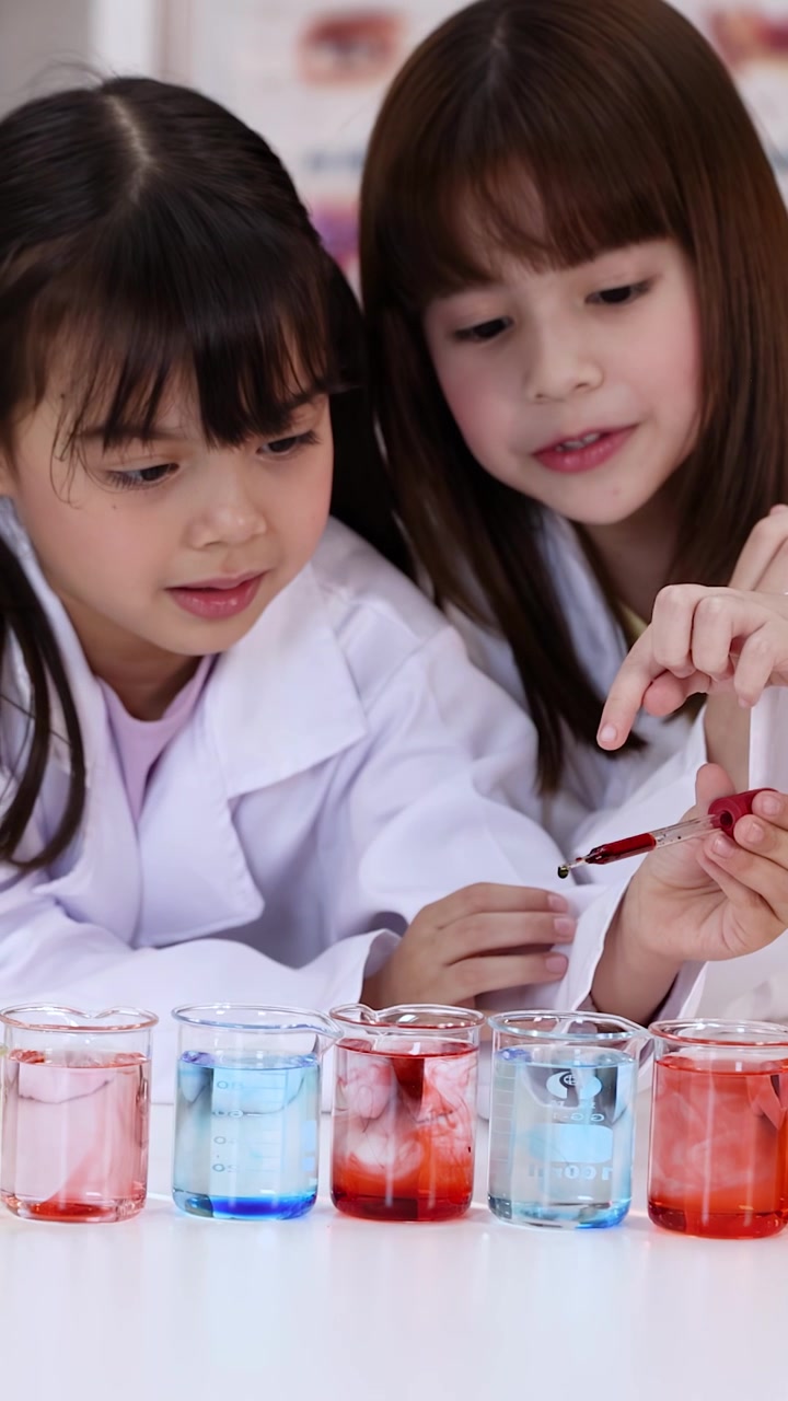 Two children in white lab coats use pipettes to drop liquid into colorful beakers.