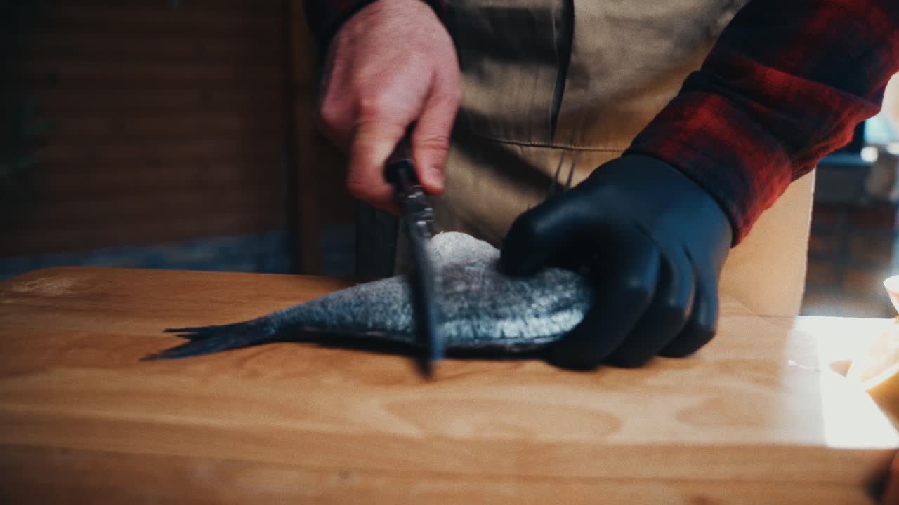 Una persona con un grembiule squama un pesce con precisione su un tagliere di legno.
