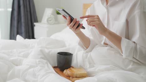 woman hands holding smartphone with social media online, coffee and bread for breakfast in the bedroom.