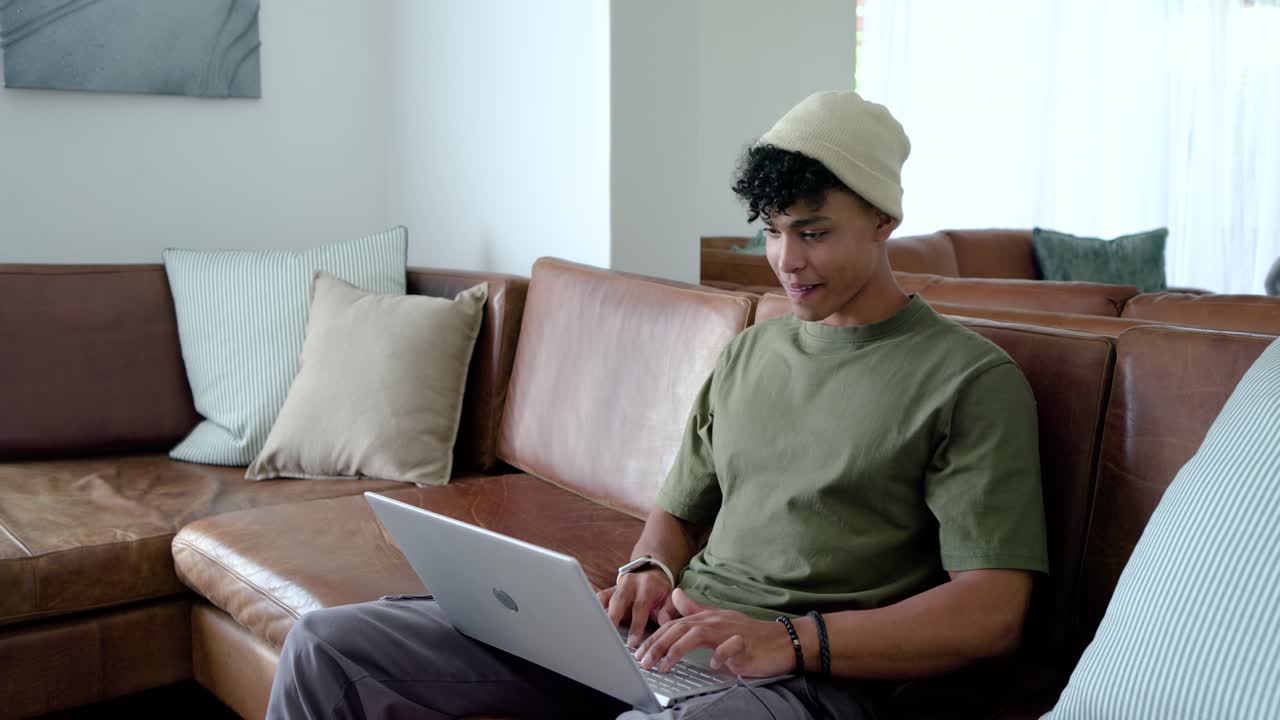 African American man using silver laptop on couch, smiling at message, leaning forward, copy space