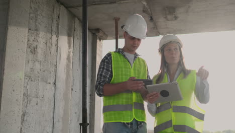 Ingenieros-Diseñadores-Un-Hombre-Y-Una-Mujer-Parados-En-El-Techo-De-Un-Edificio-En-Construcción-Y-Discuten-El-Plan-Y-El-Progreso-De-La-Construcción-Usando-Una-Tableta-Y-Un-Teléfono-Móvil.-Los-Constructores-Modernos-Discuten-La-Infraestructura-Del-Edificio-Y-Sus-Alrededores.