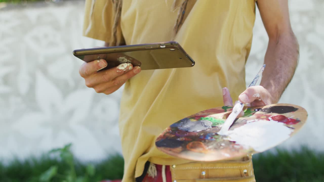Video of midsection of caucasian male artist holding tablet and painter's palette by mural on wall