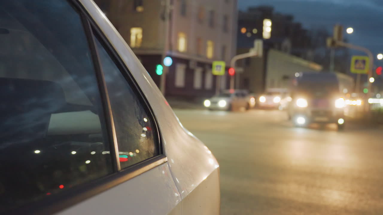 Close up side view of parked vehicle with window reflection showing city lights, while multiple cars with headlights on move along road in background under evening sky with streetlights glowing