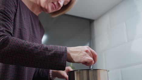 primer plano de una mujer agitando sopa en una olla de acero inoxidable en la estufa, con ropa de manga larga a cuadros, movimientos suaves de las manos, involucrada en el proceso de cocción, fondo de cocina de azulejos blancos