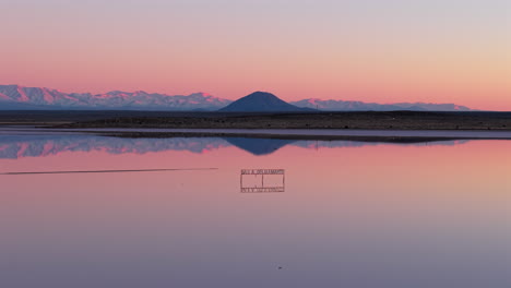 low drone pullback over calm reflective waters of salinas del diamante at sunset, showing the conical silhouette of volcán and the snowcapped andes in mendoza, argentina