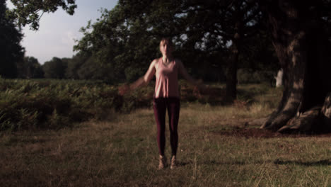 On-a-sunny-day-in-the-park-outdoors,-a-young-woman-is-seen-doing-Jumping-Jacks-in-the-lush-green-grass