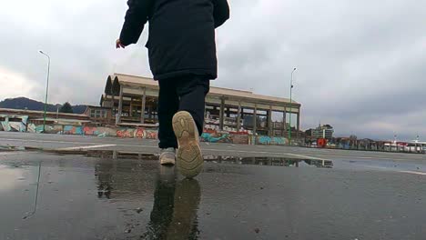 niña activa y despreocupada que sostiene una muñeca salta alto en un charco en el carril para bicicletas después de la lluvia