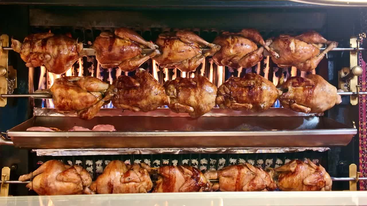 Rows of whole chickens roasting on a traditional rotisserie spit. Golden brown skin crisping over open flames at a market or butcher shop. Delicious street food cooking