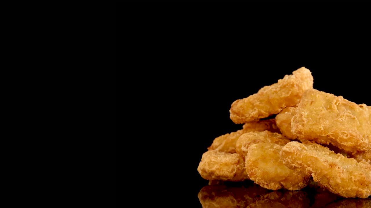 Golden, crumbed chicken nuggets rotate slowly on a glossy black surface, illuminated by dramatic studio lighting. Camera movement highlights texture and crispy coating