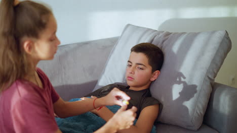 girl with ponytail and maroon shirt checks thermometer reading after removing it from sick boy resting on couch showing care concern and responsibility in calm indoor space with soft light