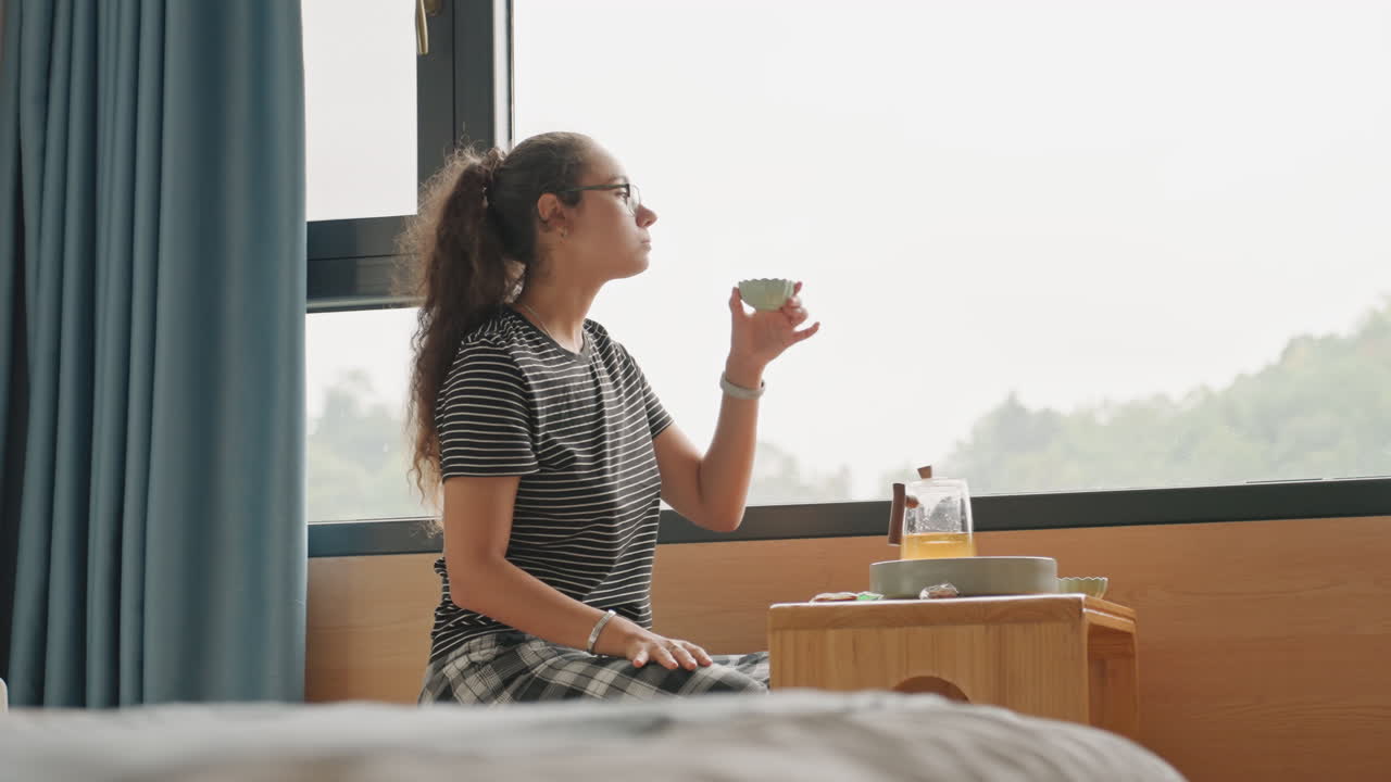 Una donna saluta l'alba. Una signora medita durante la colazione. Una donna si sofferma sull'alba dalla sua camera d'albergo. Una donna si gode la sua mattina osservando l'alba dalla finestra della sua camera d'albergo mentre sorseggia il tè.