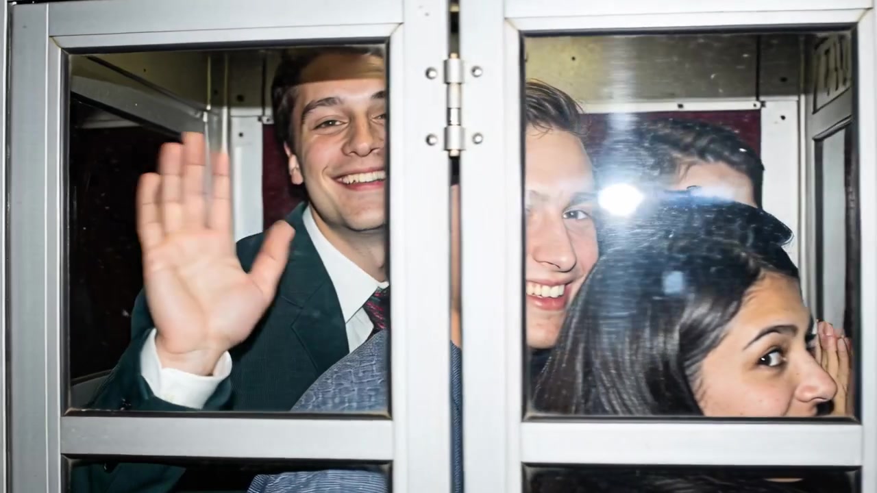 Reagendo al flash della fotocamera, cinque amici si sporgono e salutano in una cabina telefonica rossa, indossando abiti.