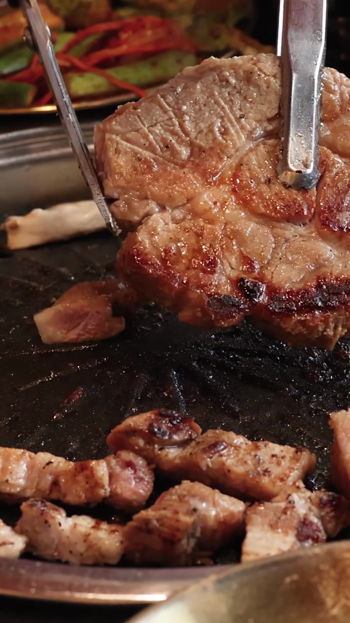 Steak babi matang diiris dengan penjepit di atas panggangan meja, pencahayaan hangat, perspektif jarak dekat.