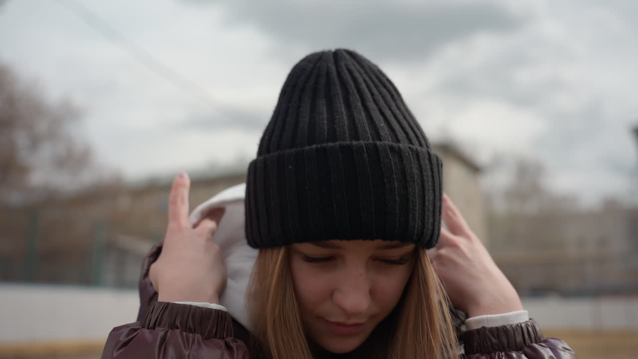 Белая женщина натягивает капюшон на черную шапку, поправляет одежду в пасмурную погоду, видна коричневая куртка и многослойная толстовка, небольшой городской ритуал укрытия и тепла перед уходом