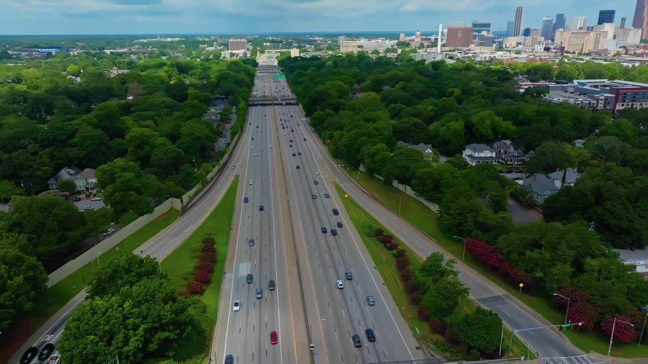 Traffic on multi lane highway in Atlanta surrounded. By green trees in summer. Aerial wide shot. Left side suburb district, right side downtown skyline. Georgia, United States