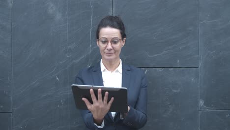 smiling caucasian businesswoman working outside, using tablet while standing at office building wall