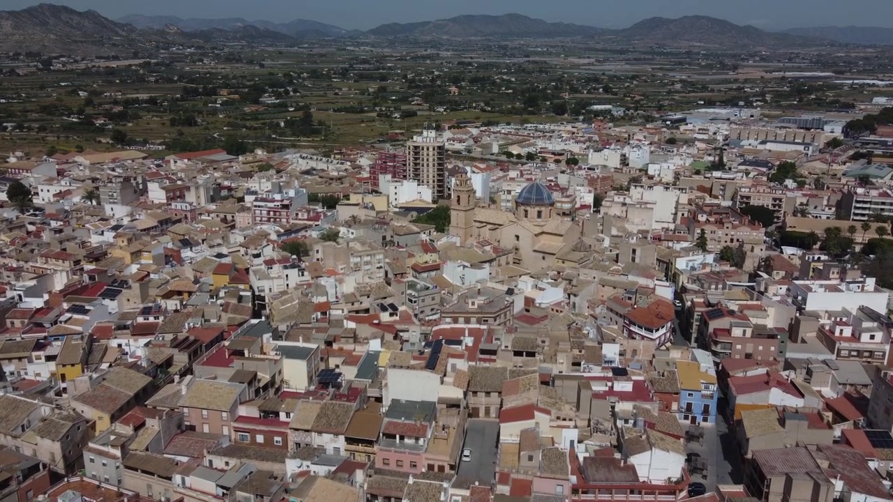 Tomada de aproximação de drone do centro histórico de Aspe, Espanha, exibindo a icônica Basílica de Nossa Senhora do Socorro. Bela arquitetura europeia, telhados de telha e layout de cidade espanhola ensolarada