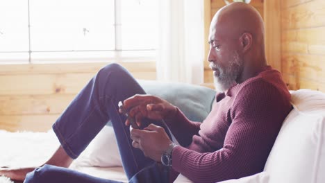 Happy-senior-african-american-man-spending-time-in-log-cabin,-sitting-on-sofa-and-using-smartphone