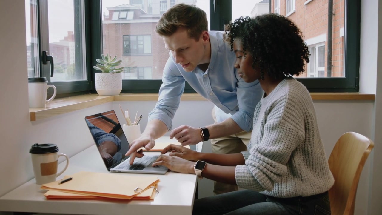 Leaning man light blue shirt pointing at laptop, woman gray sweater typing by window, guiding task