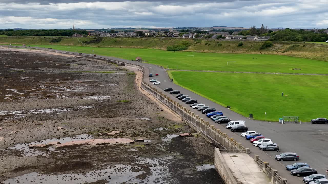Drone pans above green park, parked cars, rocky shoreline, cloudy sky, and distant townscape