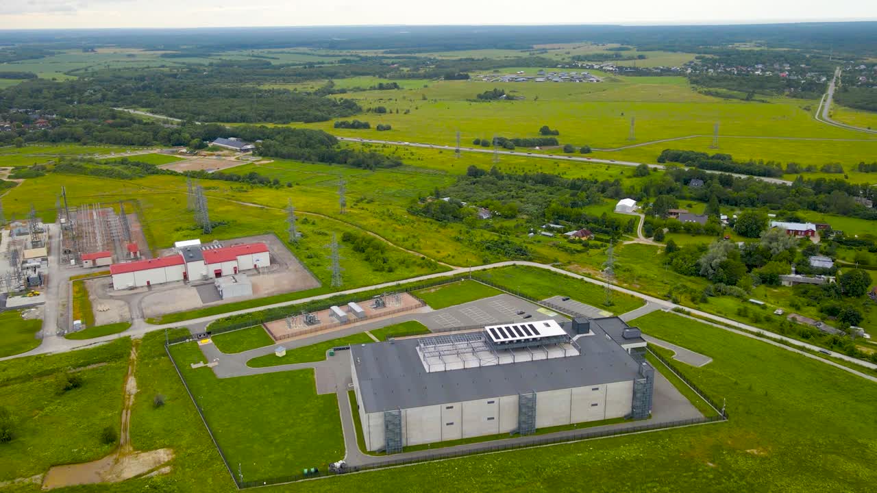 Aerial drone footage orbiting and spinning around a large modern gray colored data centre in green countryside with a large powergrid powerstation with electrical lines besides it powering the grid