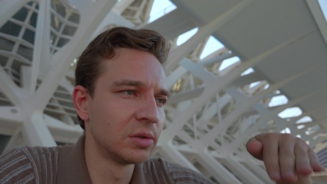 Giovane uomo siede sotto un'architettura geometrica bianca e luminosa con aperture a lucernario ripetute, passando da un profilo laterale a uno sguardo diretto, mantenendo un'espressione pensierosa e concentrata e una mano alzata.