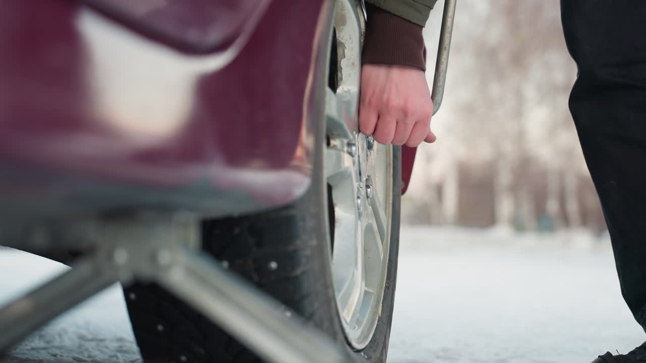 Close up of car jack positioned under vehicle on snowy ground, person walking up to car tire to loosen bolt, blurred background emphasizing tire repair action during winter maintenance