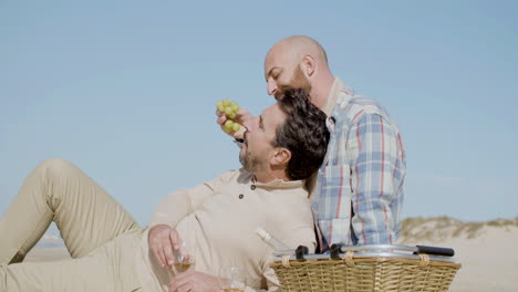 medium shot of a happy gay couple eating grape while sitting on the beach