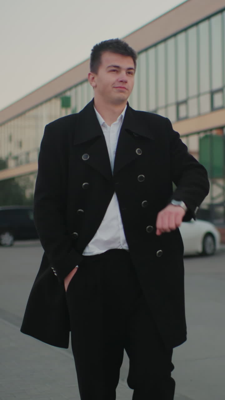 hombre con abrigo negro ajustando abrigo, poniendo la mano en el bolsillo, caminando majestuosamente con edificios de oficinas y coches estacionados en el fondo, confiado, caminando profesionalmente en un entorno de negocios urbano