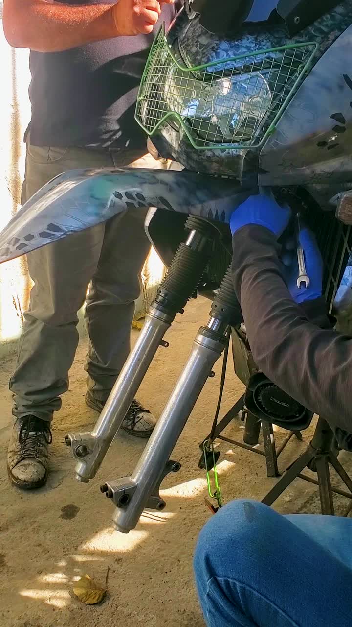 Two riders working on the front forks of Kawasaki KLR650 motorcycle. They are installing a fork brace to upgrade motorbike