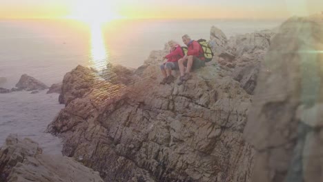 Caucasian-senior-couple-hiking-in-countryside-sitting-on-rocks-talking,-with-sunsetting-over-sea