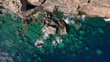 Top-down-drone-shot-of-crystal-clear-water-with-rocks-next-to-stone-cliff-and-birds-flying-by