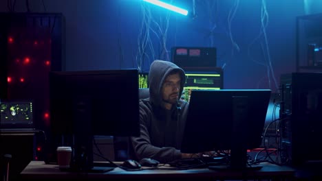 young man hacker commiting cybercrime while hacking at two computers in the dark programming room with technologies