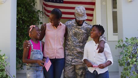 happy african american male soldier embracing wife and children in front of house and american flag