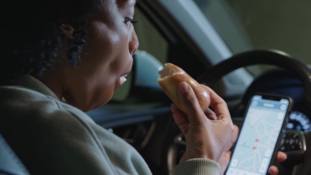 Reaching for glazed pastry after parking, driver in light sweatshirt checking directions on phone
