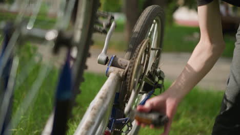 close-up partial view of someone's hand rotating bicycle pedal left, focusing on the chain and gear system, the softly blurred background includes a view of greenery and building