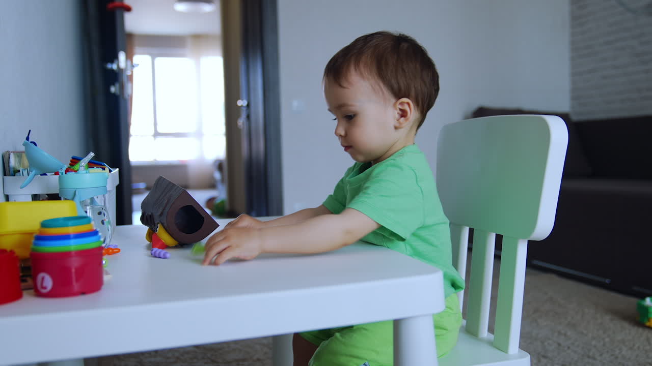 Adorable toddler boy sitting at the table. Kid plays with toys in the room. Side view.