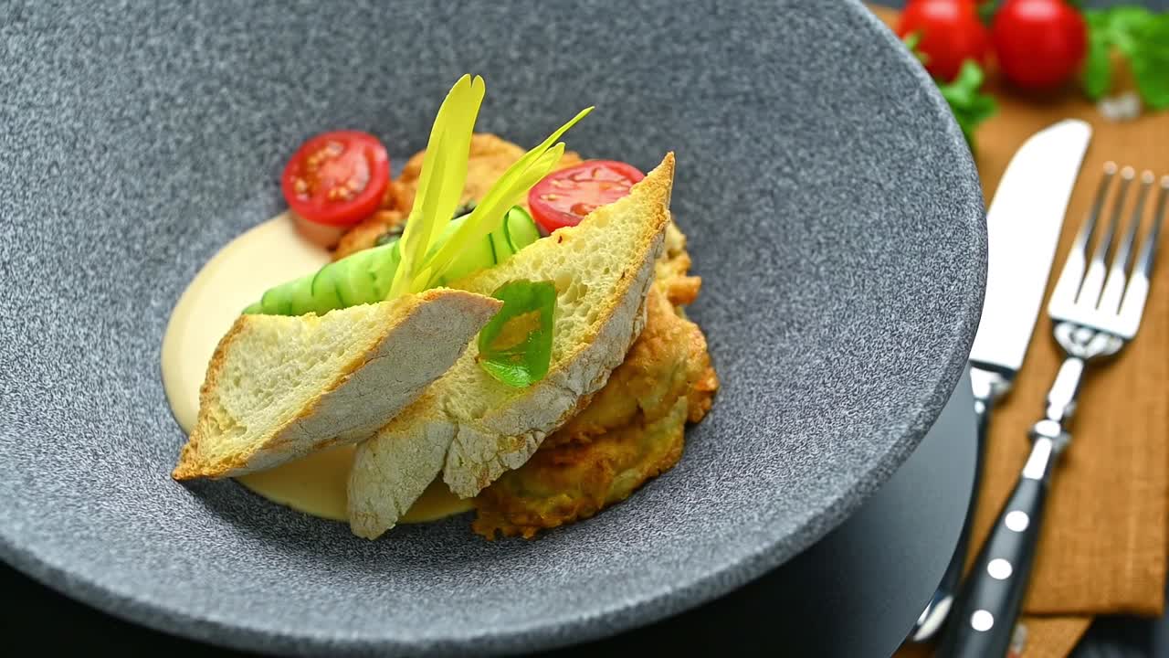 Delicioso plato servido en un plato oscuro con ingredientes frescos y decoración. La comida incluye una tortita de verduras con tomates, pepino y una salsa cremosa, todo dispuesto artísticamente.