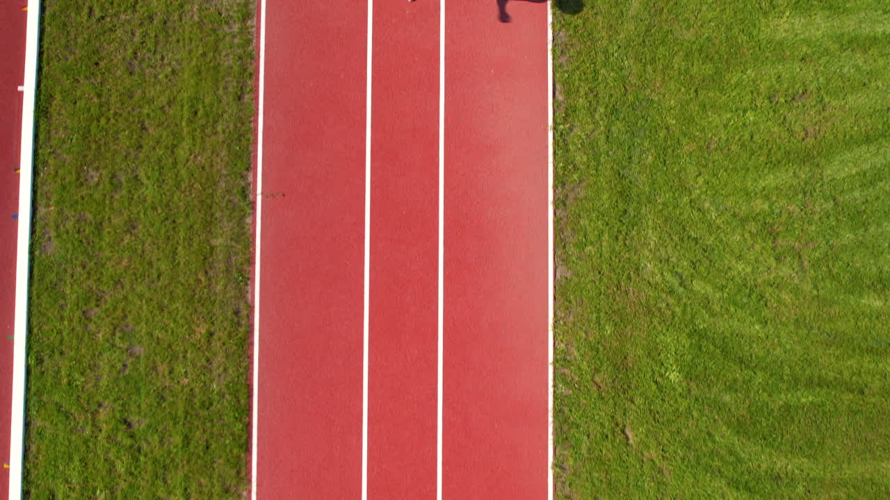 Aerial view of an athlete running on a red track, with grass on both sides and a long shadow cast on the track surface under bright sunlight.