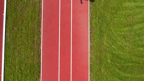 aerial view of an athlete running on a red track, with grass on both sides and a long shadow cast on the track surface under bright sunlight.
