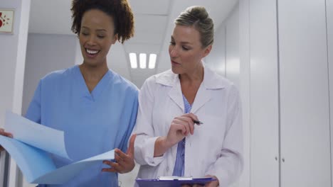 diverse female doctor and medical worker standing in hospital corridor and talking