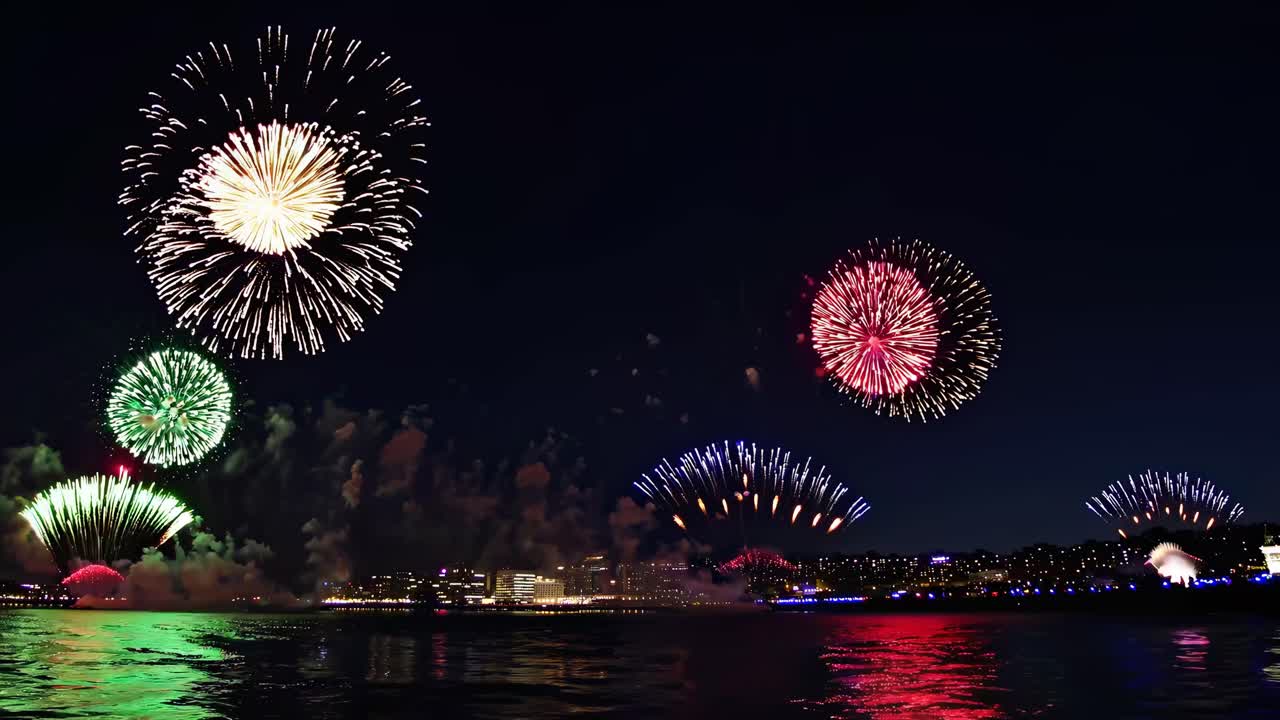 Vibrant fireworks explode over a city skyline at night, captured from a low-angle video