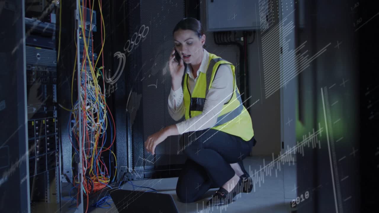 Receiving call female tech kneeling in rack aisle using laptop pointing cables wearing vest fixing