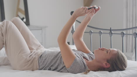 woman wearing pyjamas posing and taking selfies on mobile phone lying on bed