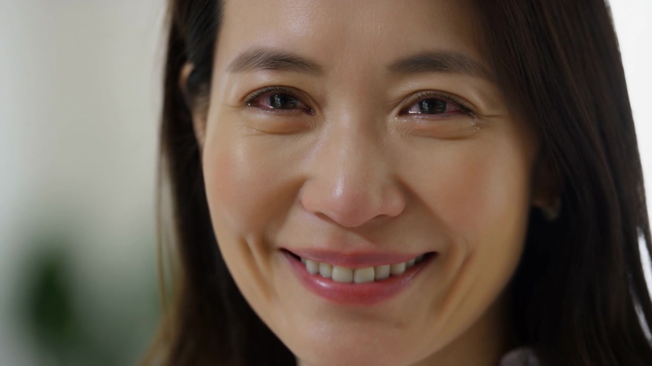 Close view shows an adult woman maintaining eye contact and a gentle smile, with subtle facial movement, soft natural-looking light, and an out-of-focus interior behind her.