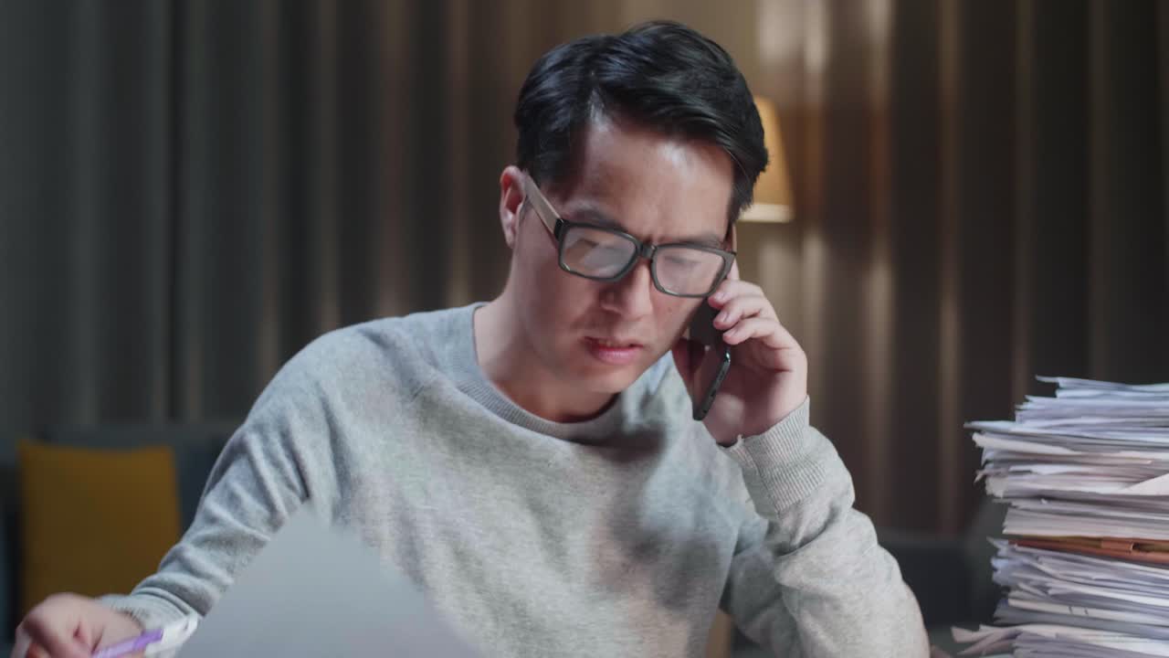 집에서 문서와 함께 열심히 일하는 동안 전화로 이야기하고 메모를 취하는 아시아 남자의 클로즈업