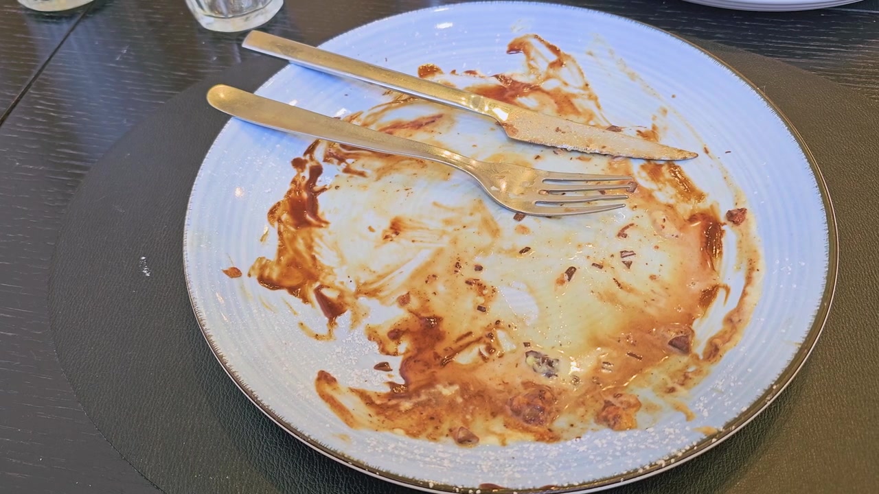 Finished cafe dessert plate with dirty fork and knife, chocolate sauce streaks, waffle crumbs, powdered sugar, and cream residue on a blue plate after a sweet brunch meal indoors close up.