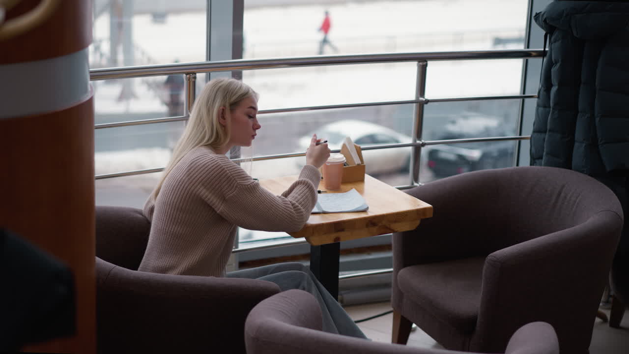 joven sentada pensativa, continuando escribiendo con dos asientos vacíos y chaqueta colgada, vista borrosa de personas caminando y coches estacionados en el fondo de un acogedor café