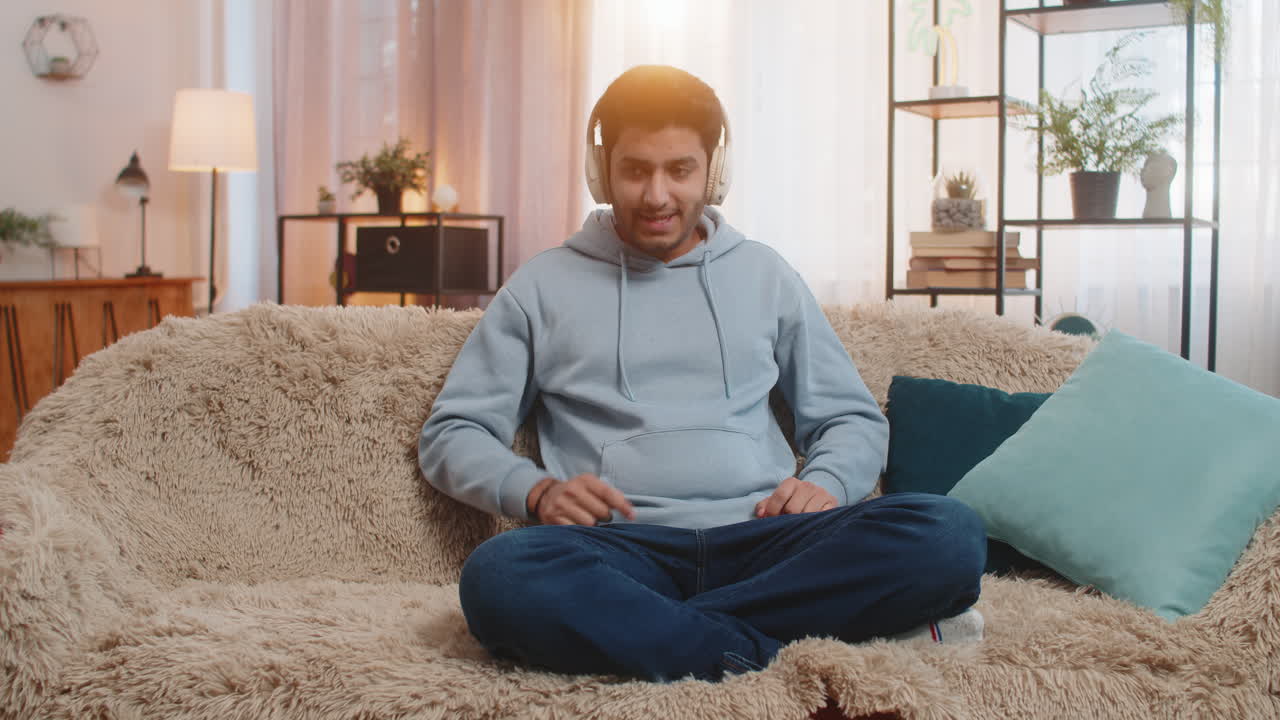 Indian young man sitting on sofa listening to music with headphones dancing to rhythm at home