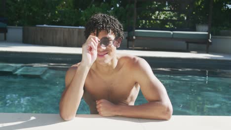 retrato de un hombre biracial feliz con gafas de sol en la piscina en el jardín en un día soleado.